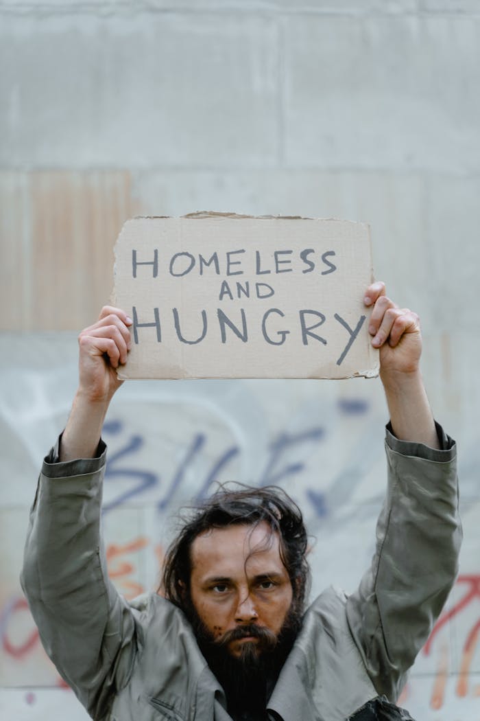 A bearded homeless man holds a sign reading 'Homeless and Hungry' against a graffiti wall.